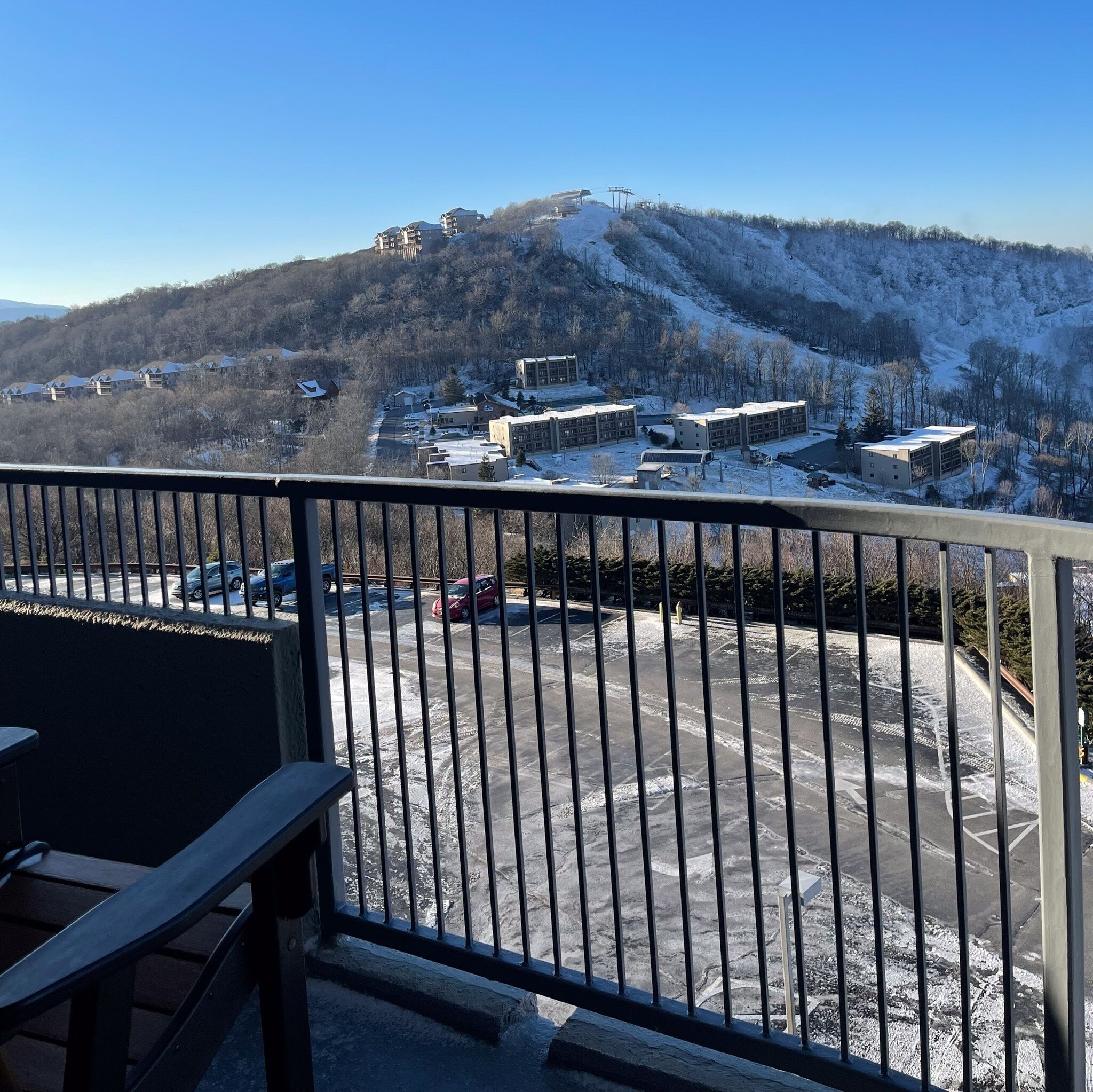 Blick vom Balkon auf einen Parkplatz und Gebäude an einem verschneiten Hang.