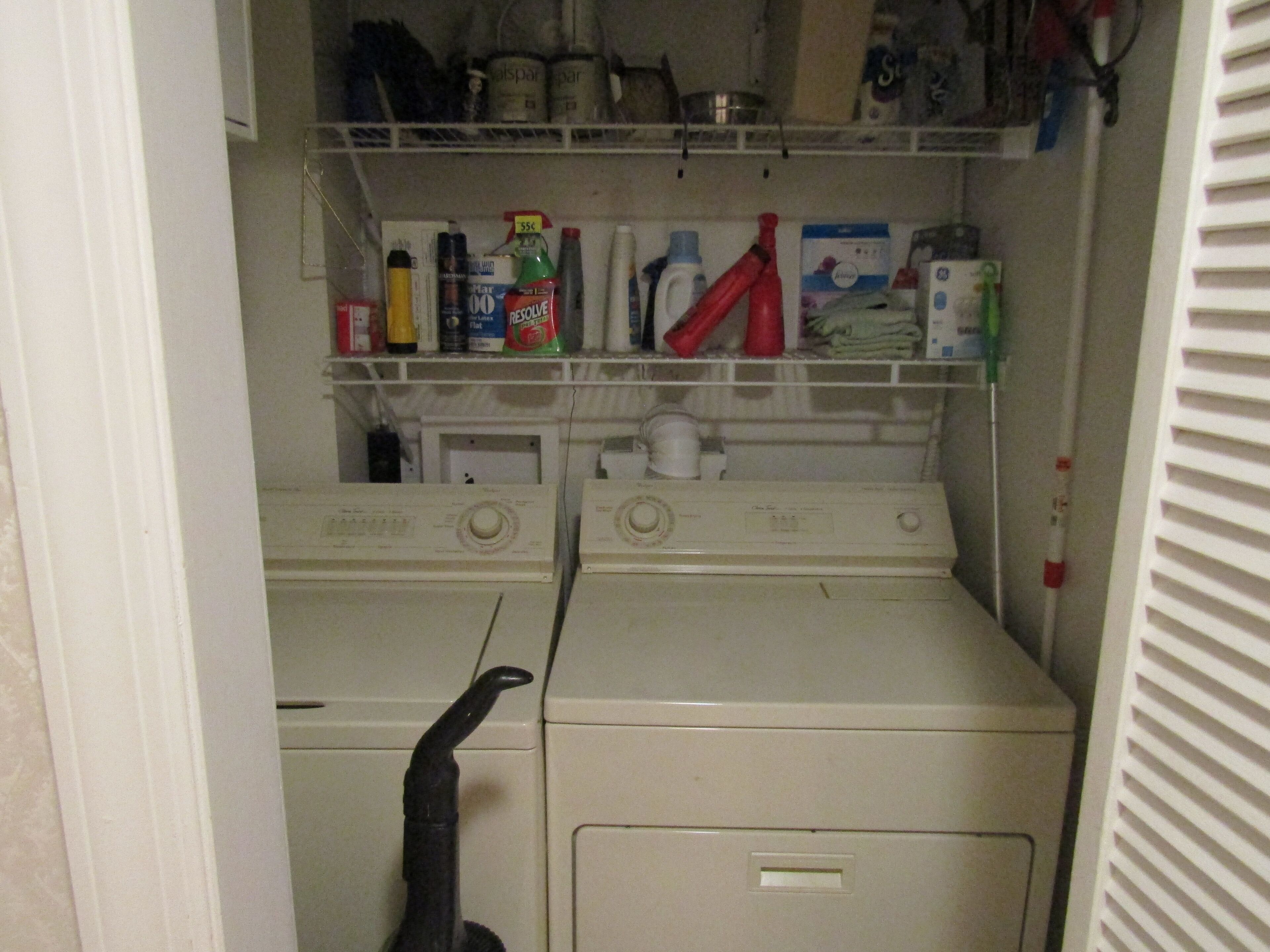 A laundry room with two washing machines, a dryer, and shelves above containing cleaning supplies.