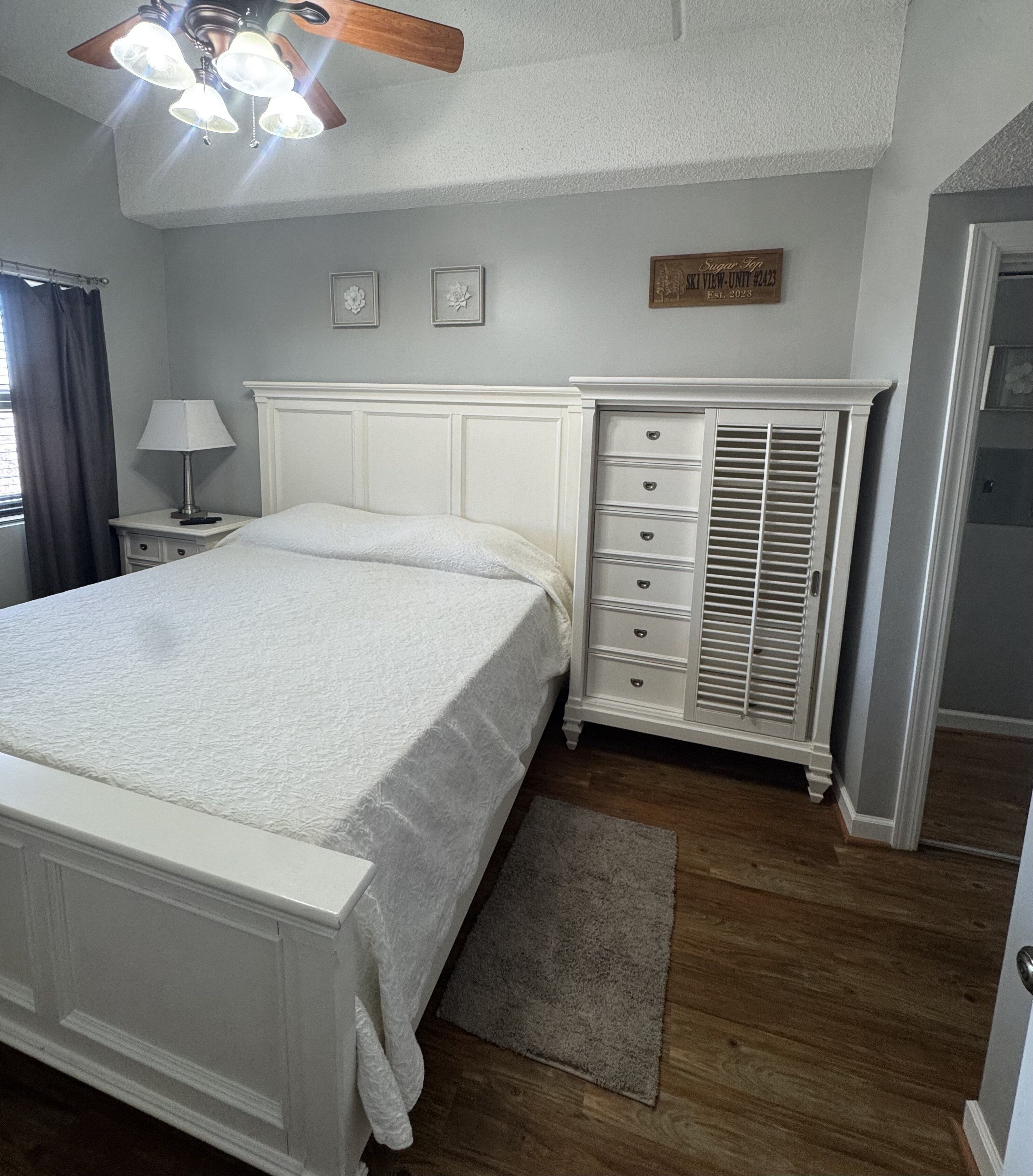 A bedroom with a bed, a dresser, a nightstand, a ceiling fan, and a door.