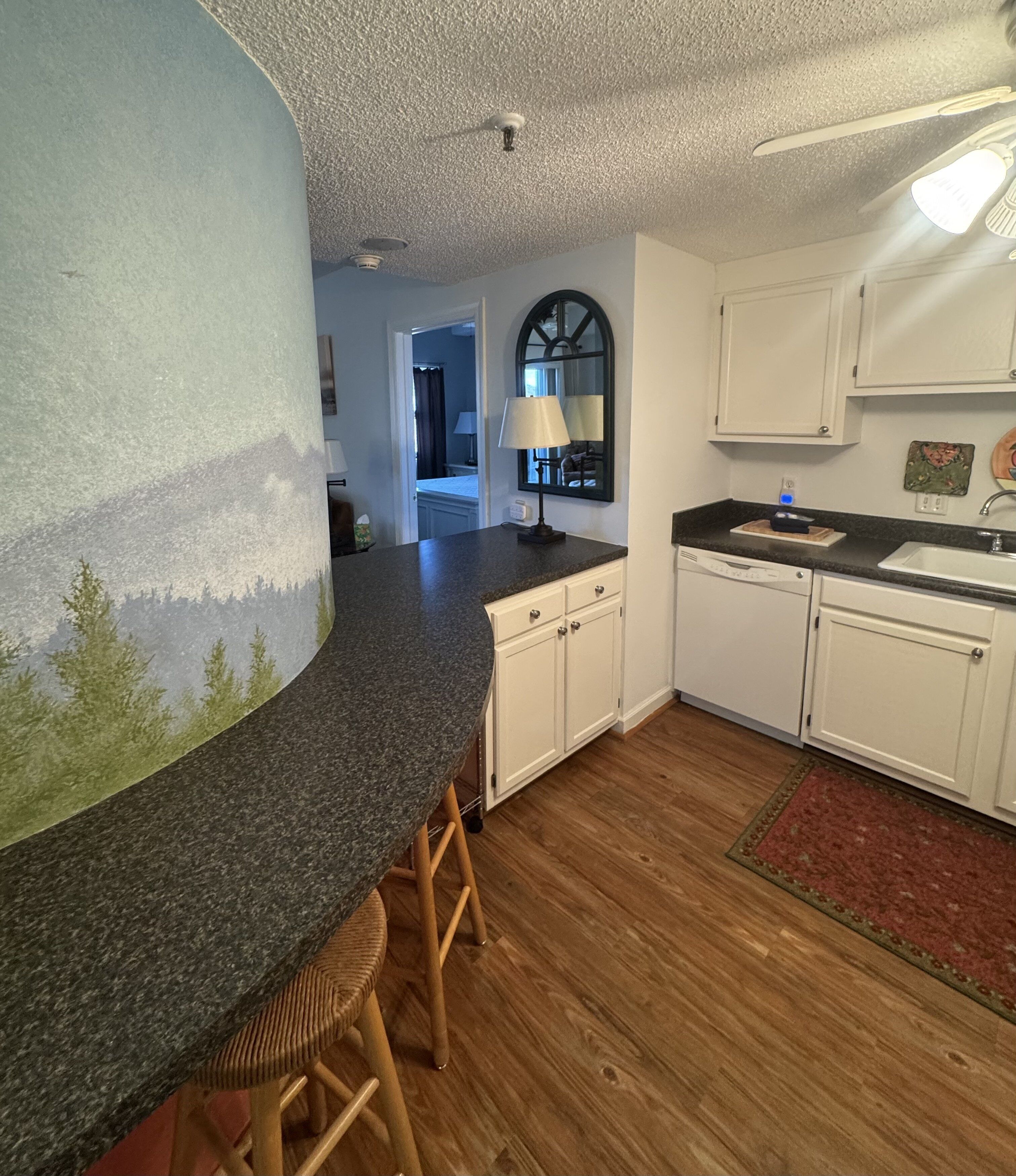 A kitchen with a curved granite countertop, wooden stools, white cabinets, and a red rug.