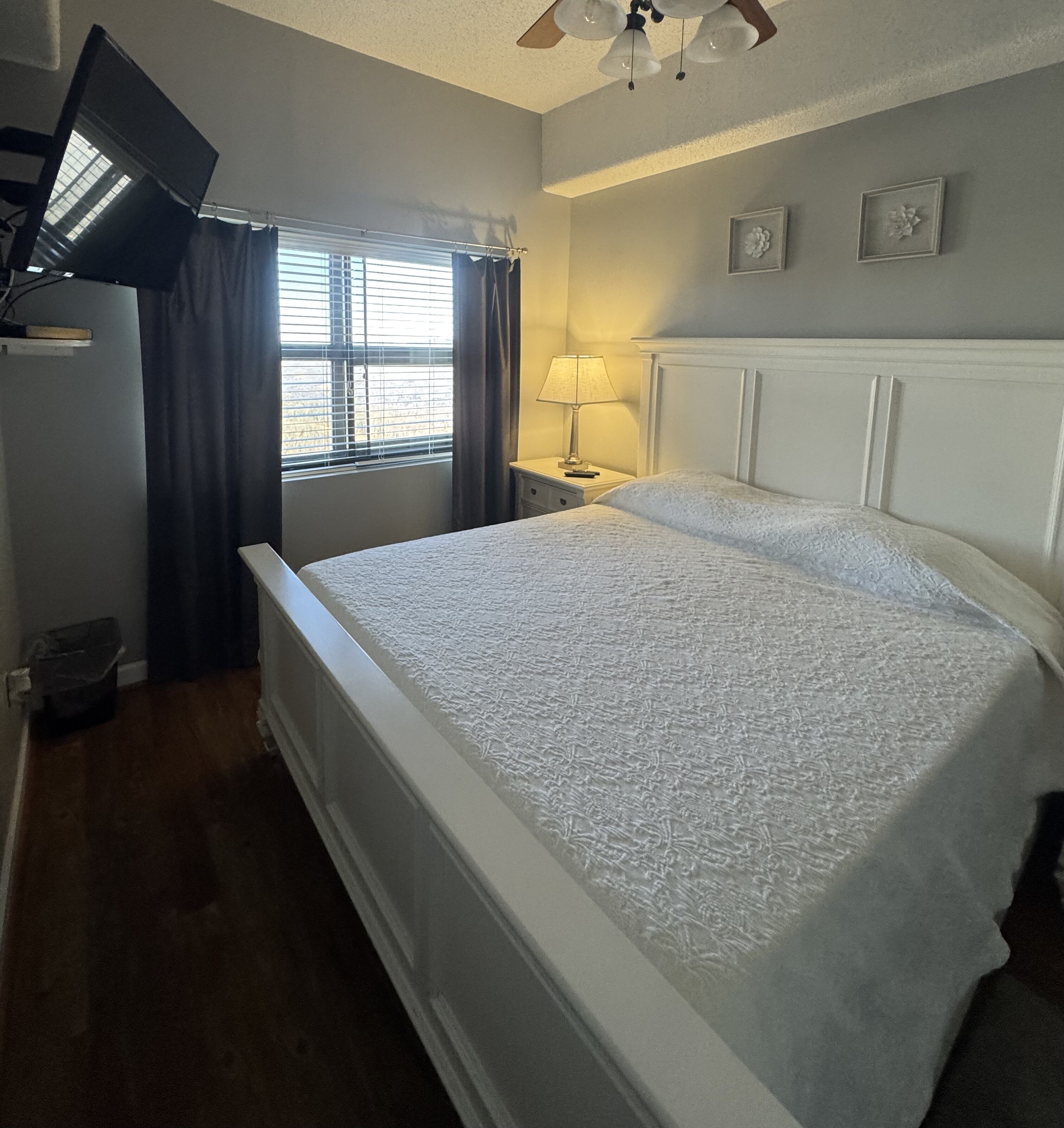 Une chambre bien rang ée, avec un lit, une table de chevet, une lampe, un téléviseur fixé au mur et une fenêtre avec des rideaux.