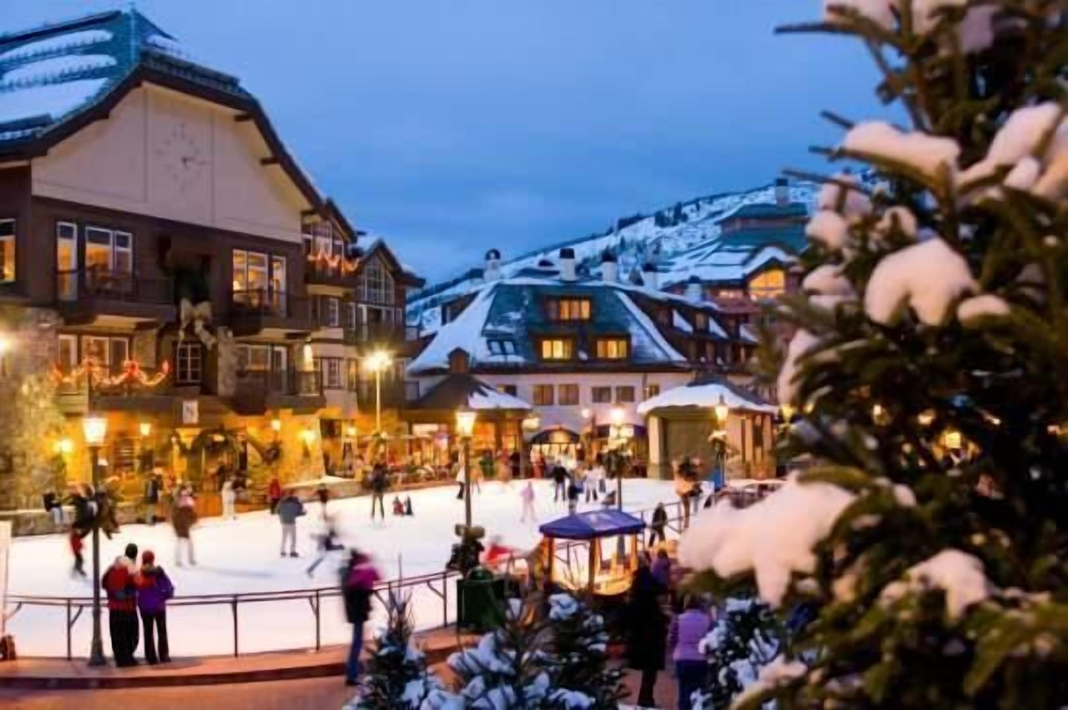 Une patinoire en plein air recouverte de neige, entourée de bâtiments traditionnels illuminés d’une lumière chaleureuse.
