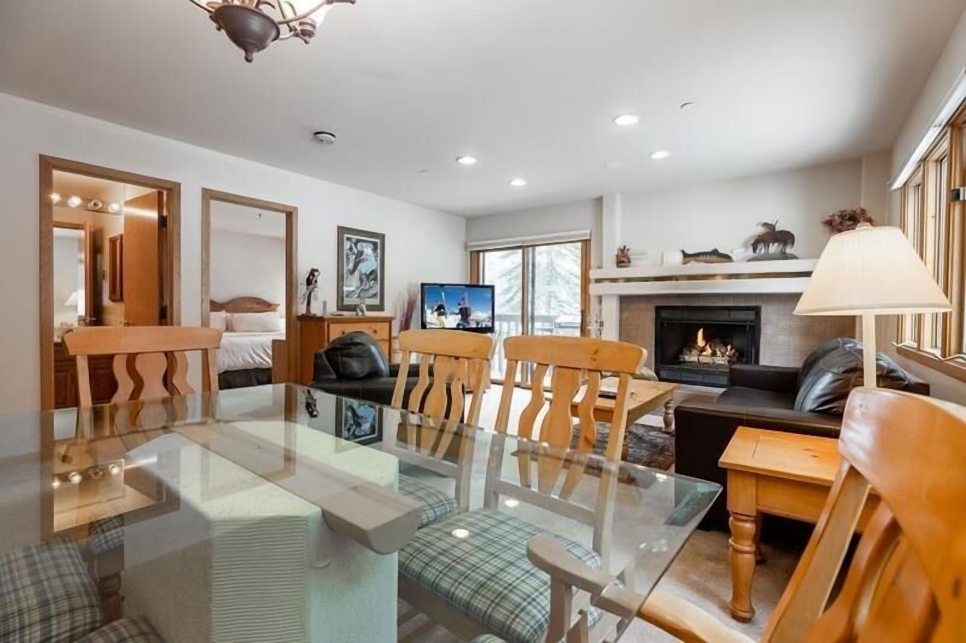 A modern living room with a glass-top dining table, wooden chairs, a fireplace, and a television.