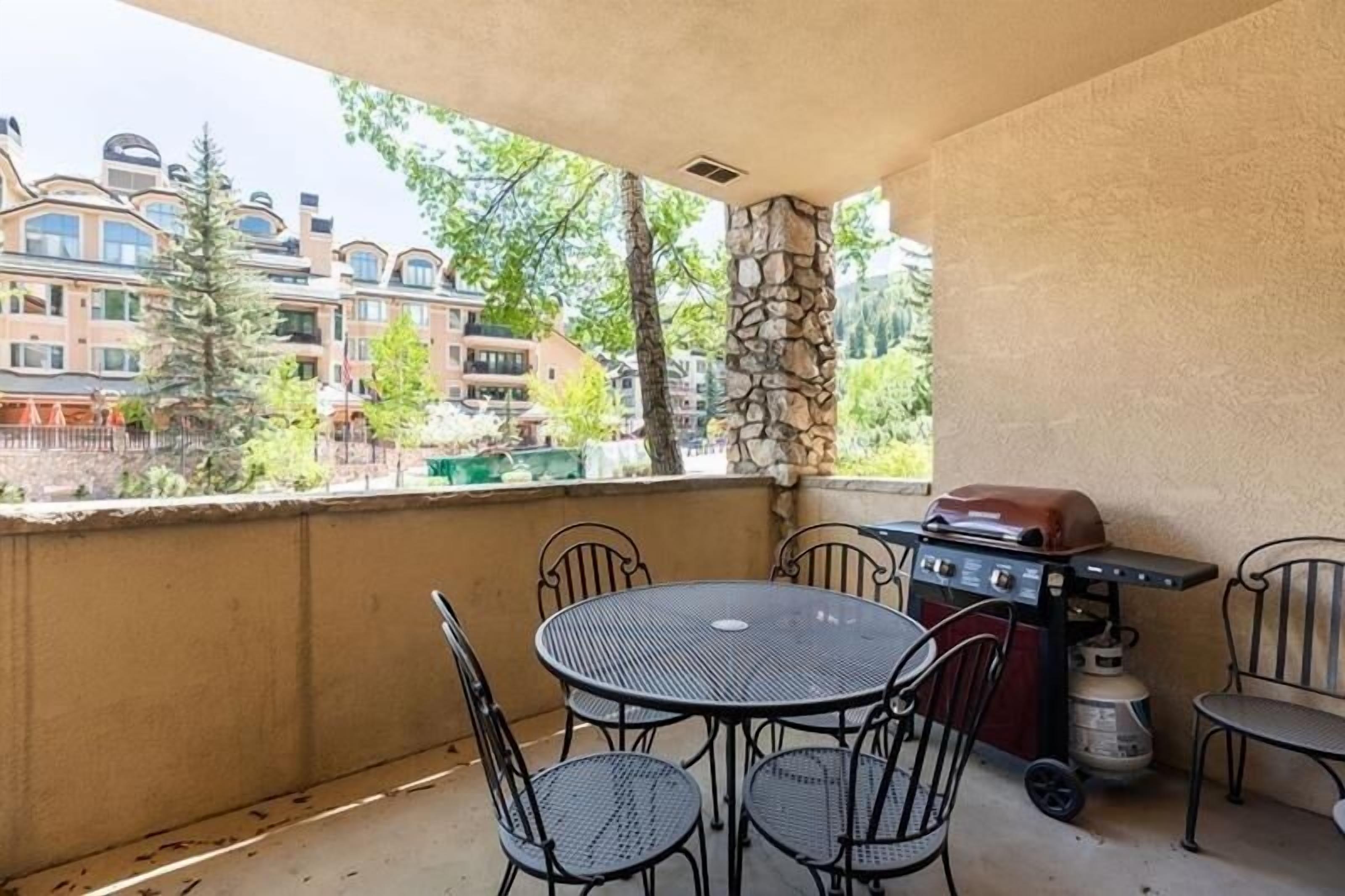 A covered outdoor patio with a round metal table and four chairs, a barbecue grill, and a propane tank.