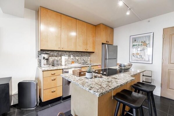 A modern kitchen with wooden cabinets, a granite countertop, a stainless steel refrigerator, and a small island with stools. - Vail Ski Resort - Luxury Condos (Vail)