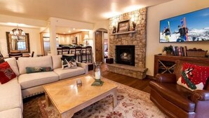 A cozy living room with a stone fireplace, a television mounted above it, and a dining area in the background. - Vail Ski Resort - Luxury Condos (Vail)
