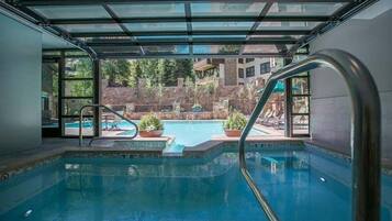 An indoor swimming pool with clear glass walls, a poolside area with lounge chairs, and a view of a landscaped garden and buildings in the background.
