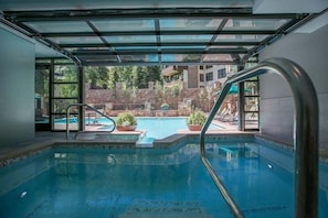 En inomhuspool med väggar av genomskinligt glas, ett område vid poolen med solstolar och utsikt över en vackert anlagd trädgård och byggnader i bakgrunden.