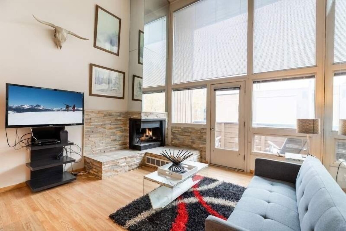 A modern living room with a fireplace, large windows, and a mounted TV.