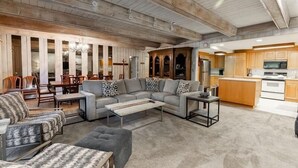Ein geräumiger Wohnbereich mit Sofa, Hocker und Couchtisch, Holzbalken an der Decke sowie eine Küche mit Holzschränken und Geräten.