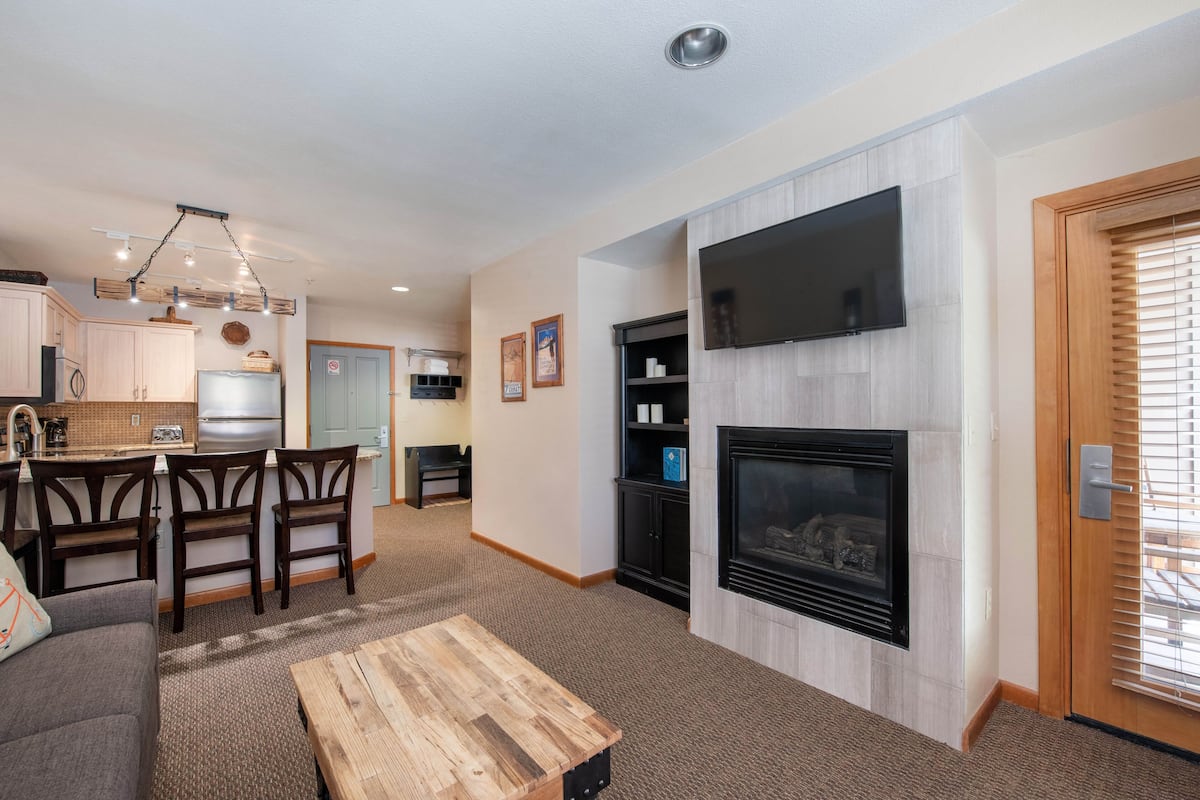 A modern living room with a fireplace, a TV mounted on the wall, and a dining area with wooden chairs and a table.