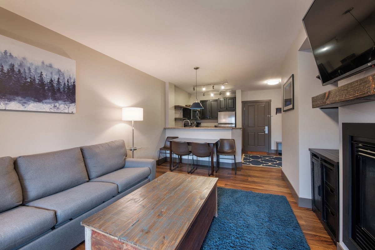 A modern living room with a gray sofa, a wooden coffee table, and a blue rug. A kitchen with dark cabinets and a dining area with chairs is visible in the background.