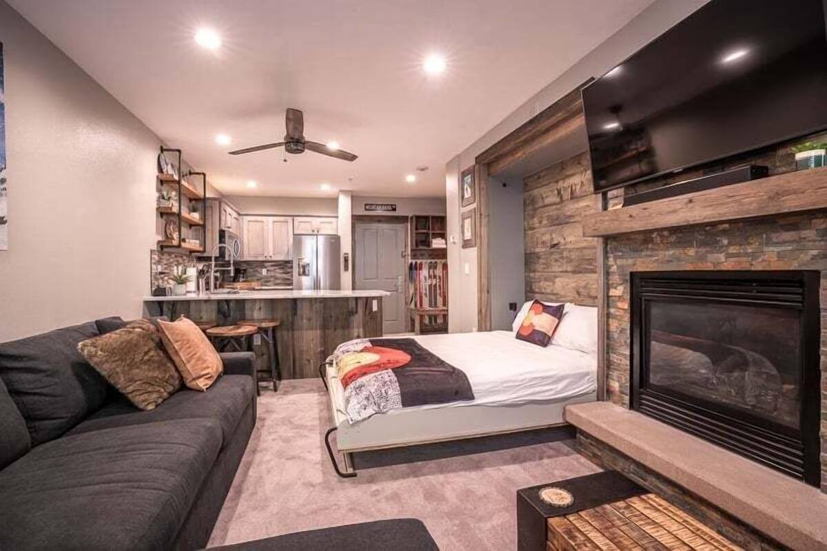 A modern living room with a sofa, a bed, a fireplace, and a kitchen in the background.