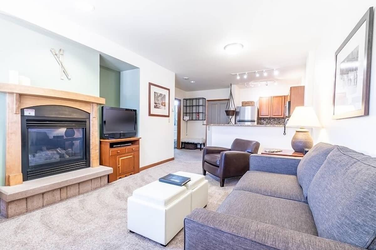 A living room with a fireplace, a sofa, a television, and a kitchen in the background.
