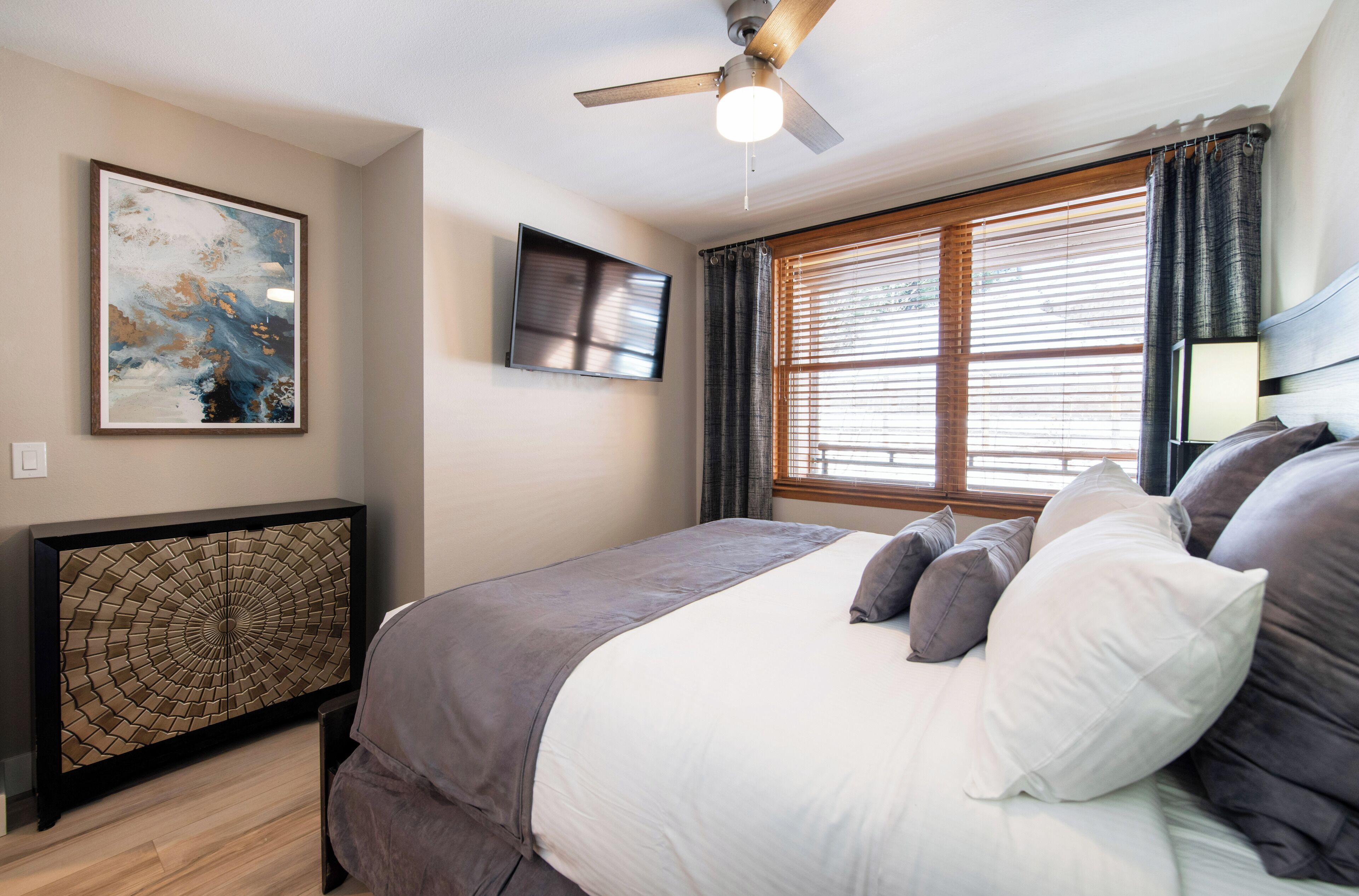 Un dormitorio con cama, ventilador de techo, televisor y ventana con cortinas.