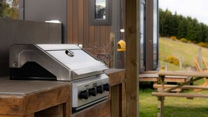 Een roestvrijstalen barbecue met vier bedieningsknoppen, geplaatst op een houten tafel voor een modern huis met een raam en uitzicht op een gazon.