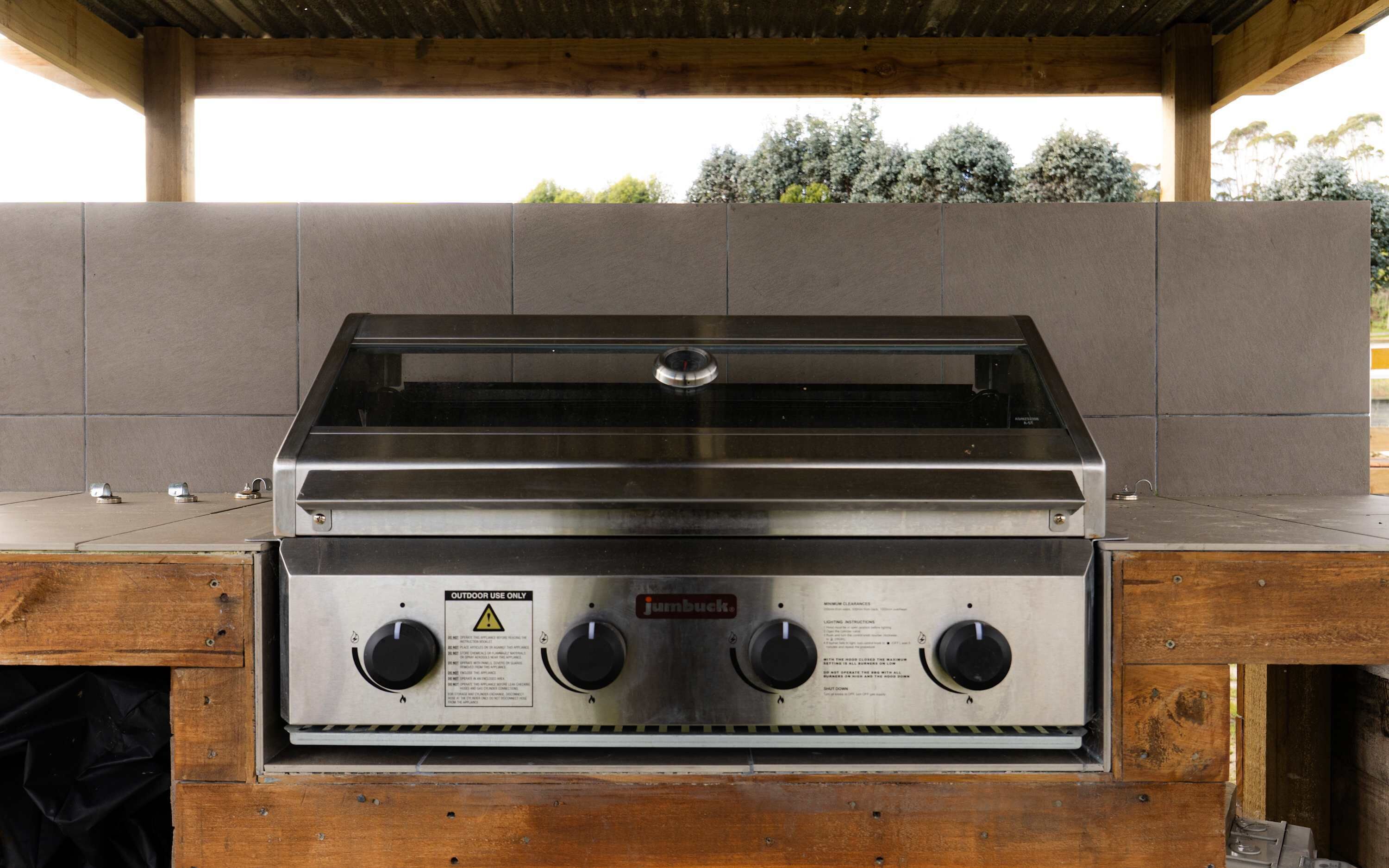 A stainless steel outdoor grill with a wooden base, labeled for outdoor use, under a wooden pergola.