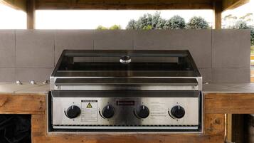 A stainless steel outdoor grill with a wooden base, labeled for outdoor use, under a wooden pergola.