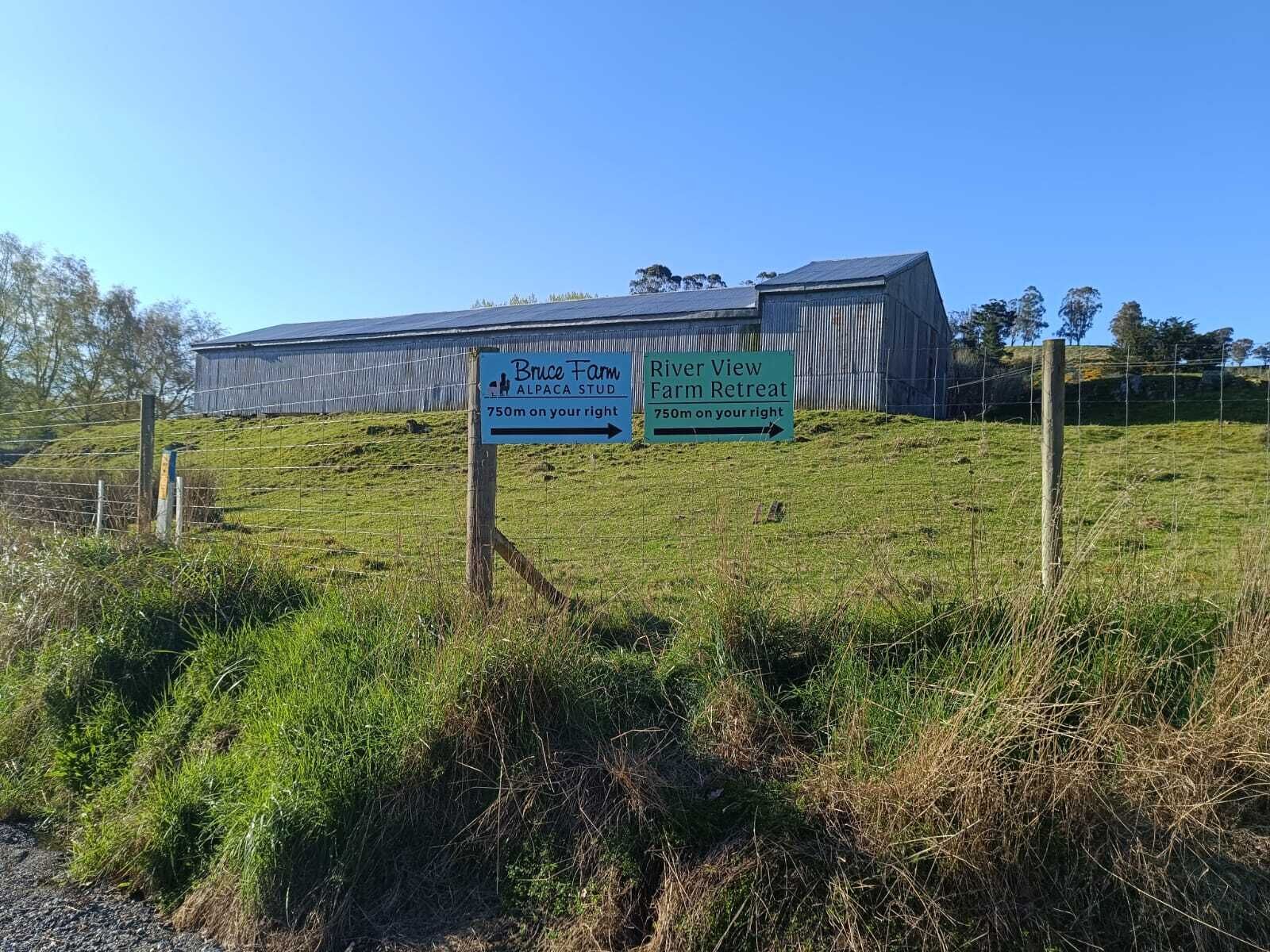 Paesaggio rurale con un edificio agricolo e due segnali stradali che indicano 'Bruce Farm Alpaca Stud' e 'River View Farm Retreat' a 750 metri sulla destra.