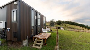 Eine moderne, dunkel gefärbte Holzhütte mit einer großen Schiebetür, einer kleinen Sitzgelegenheit im Freien mit zwei weißen Stühlen und einem Holztisch.
