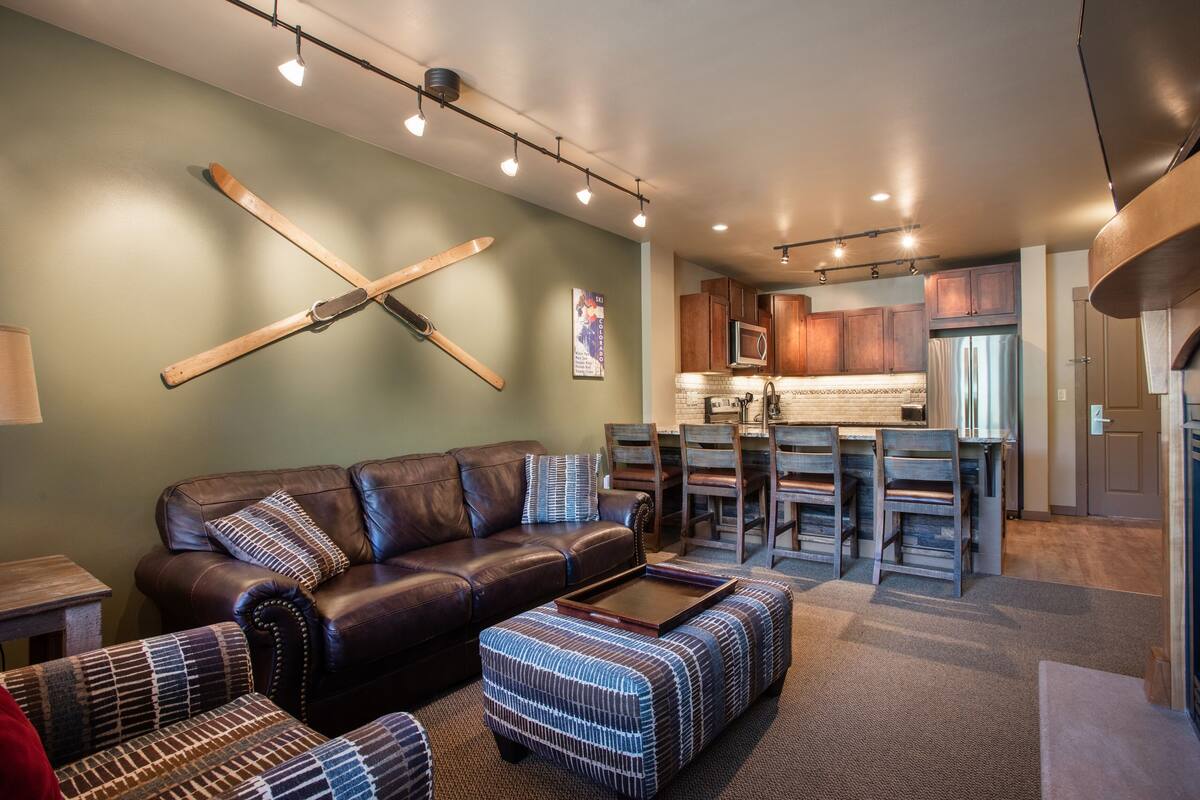 A modern living room with a leather sofa, patterned ottoman, and a dining area with wooden chairs and a bar.