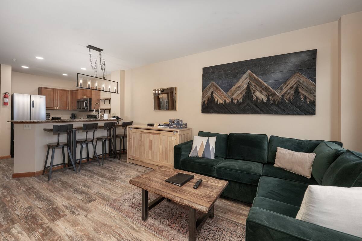 A modern living room with a green sofa, a coffee table, and a kitchen area with a bar and stools.