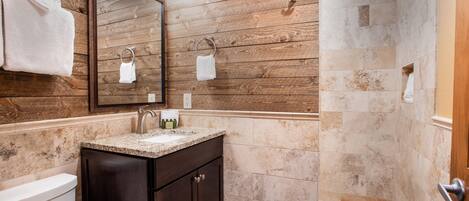 Un baño con pared de madera, un lavabo con encimera de mármol, armarios oscuros, inodoro y zona de ducha.