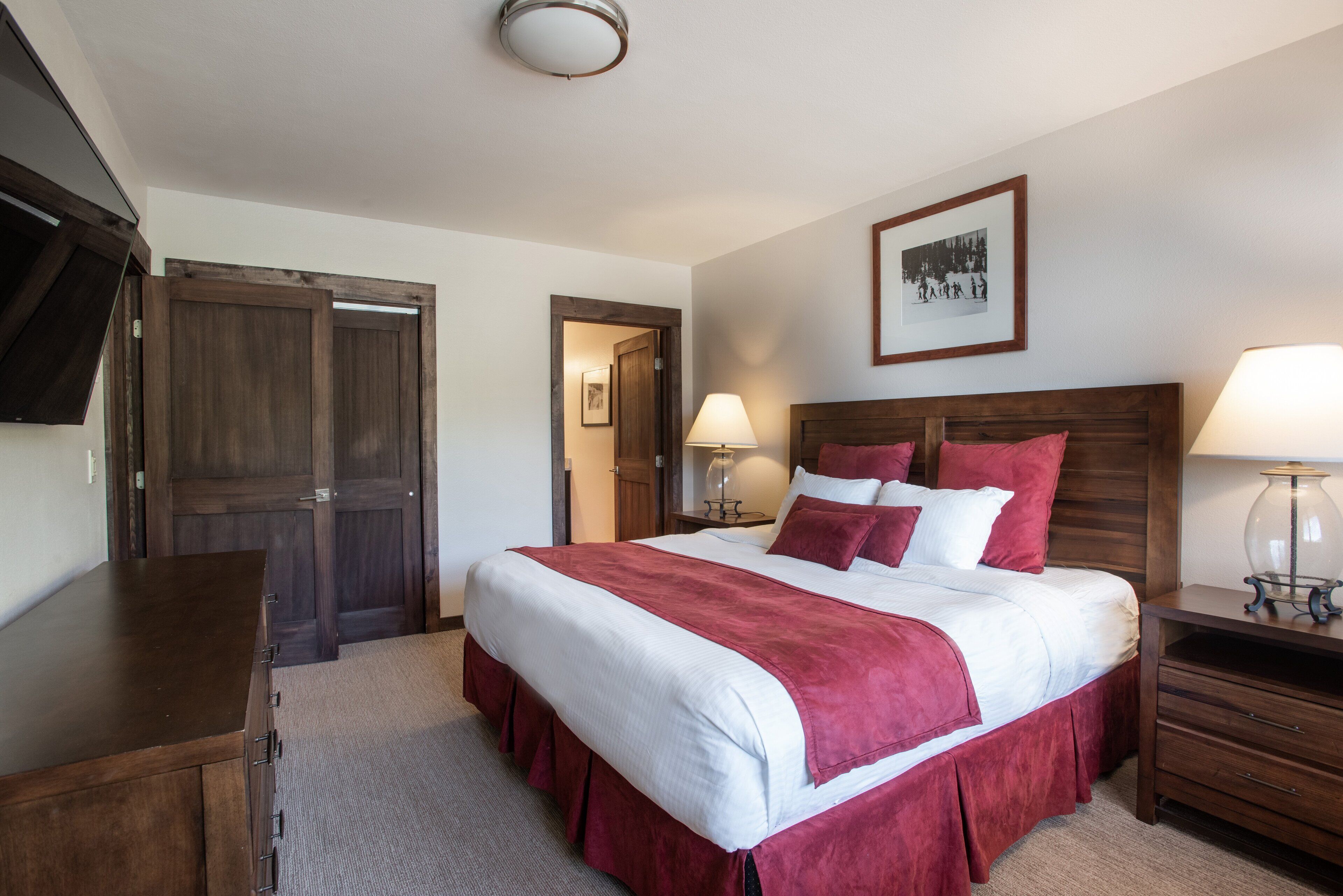A hotel room with a large bed, wooden headboard, a flat-screen TV mounted on the wall, a bedside table with a lamp, and a door leading to another room.