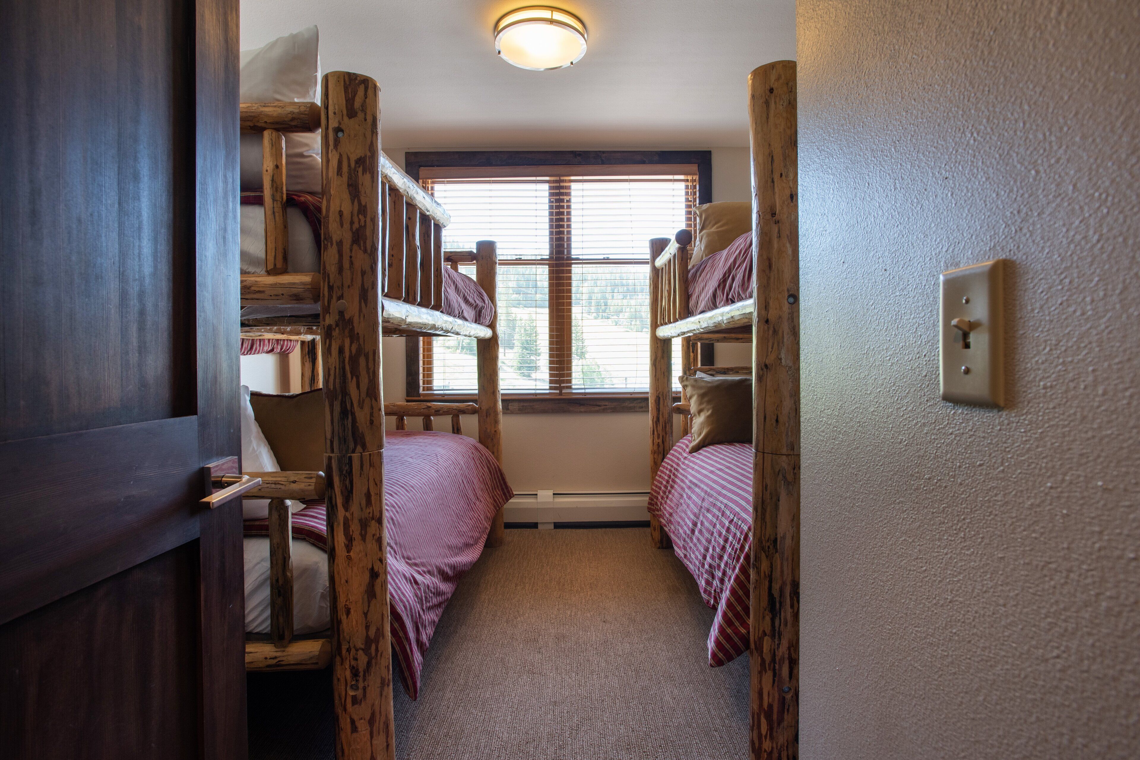 Ein schmales Zimmer mit Etagenbetten, einem Fenster mit Jalousien und einem Lichtschalter an der Wand.