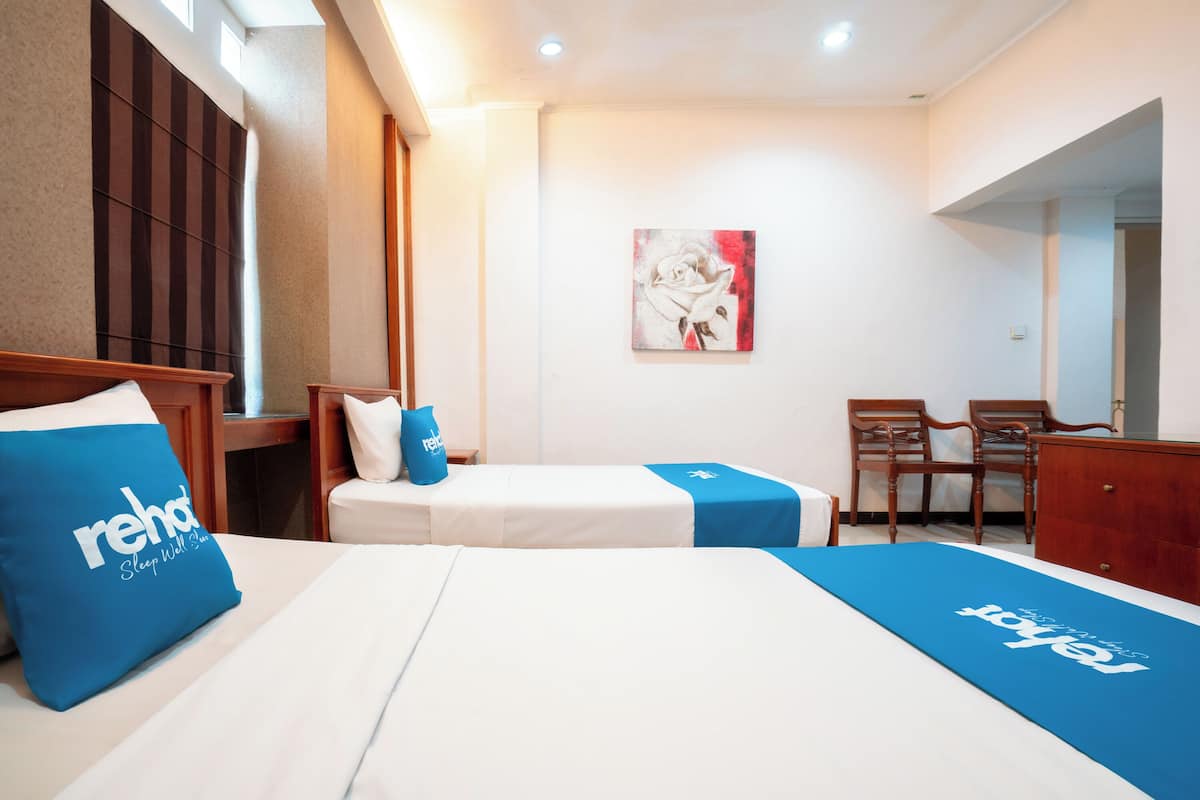 A hotel room with a bed, a desk, and a chair. There is a painting on the wall and a blue pillow with text on it.