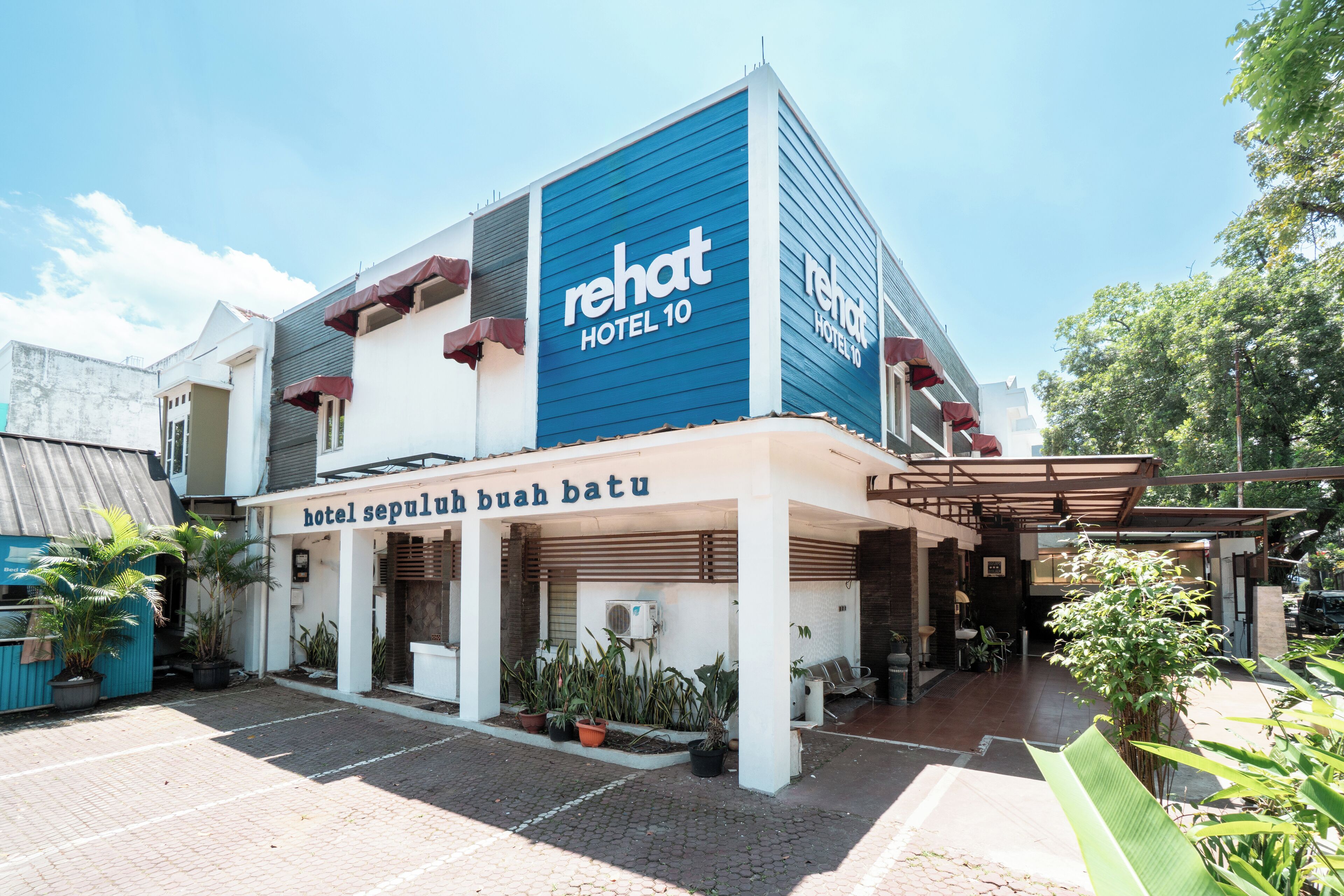 A hotel with a blue facade, a sign reading 'Rehat Hotel 10', and a covered entrance area with potted plants.