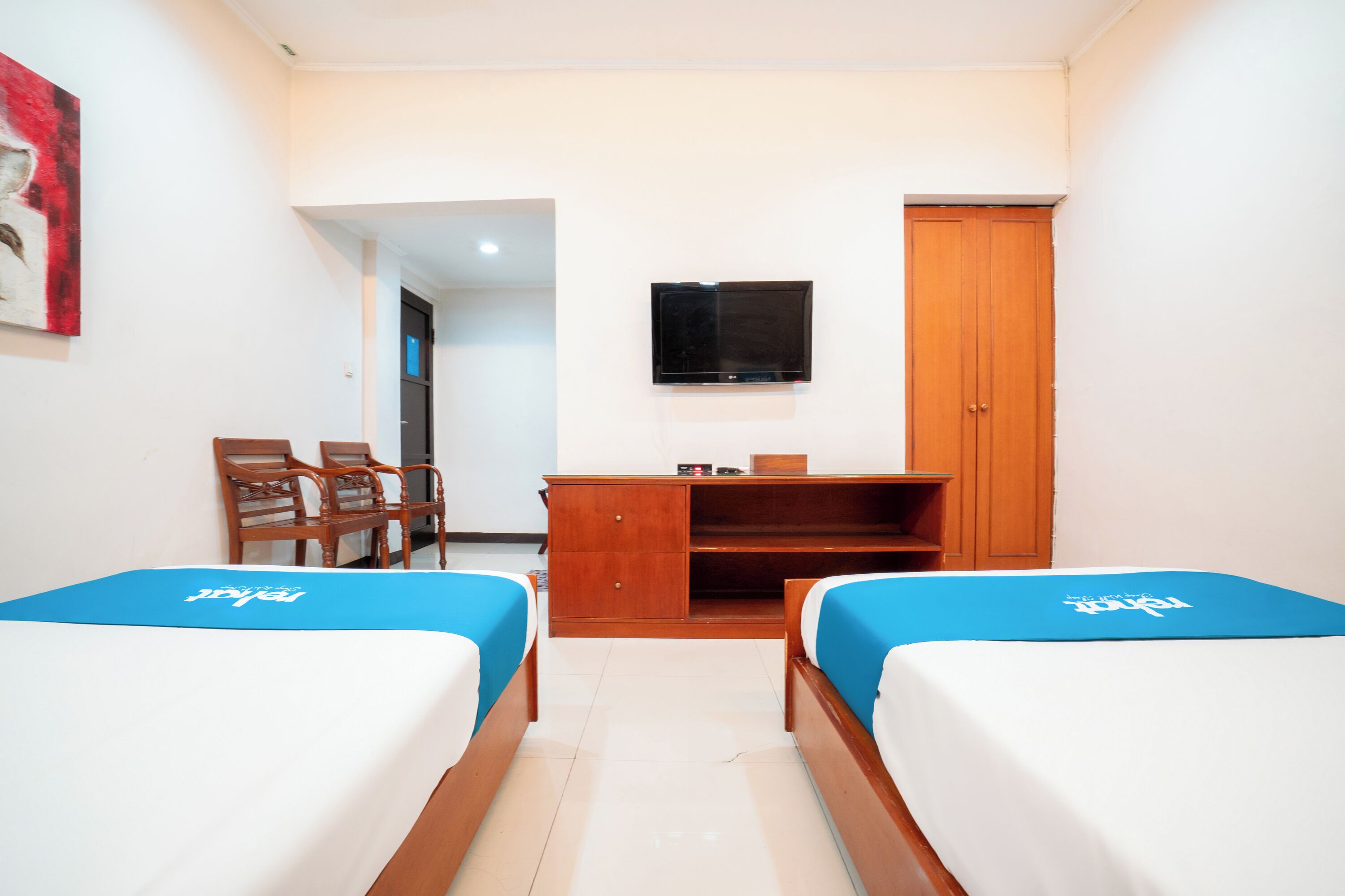 A hotel room with two single beds, a wooden desk, a TV mounted on the wall, and a wooden wardrobe.