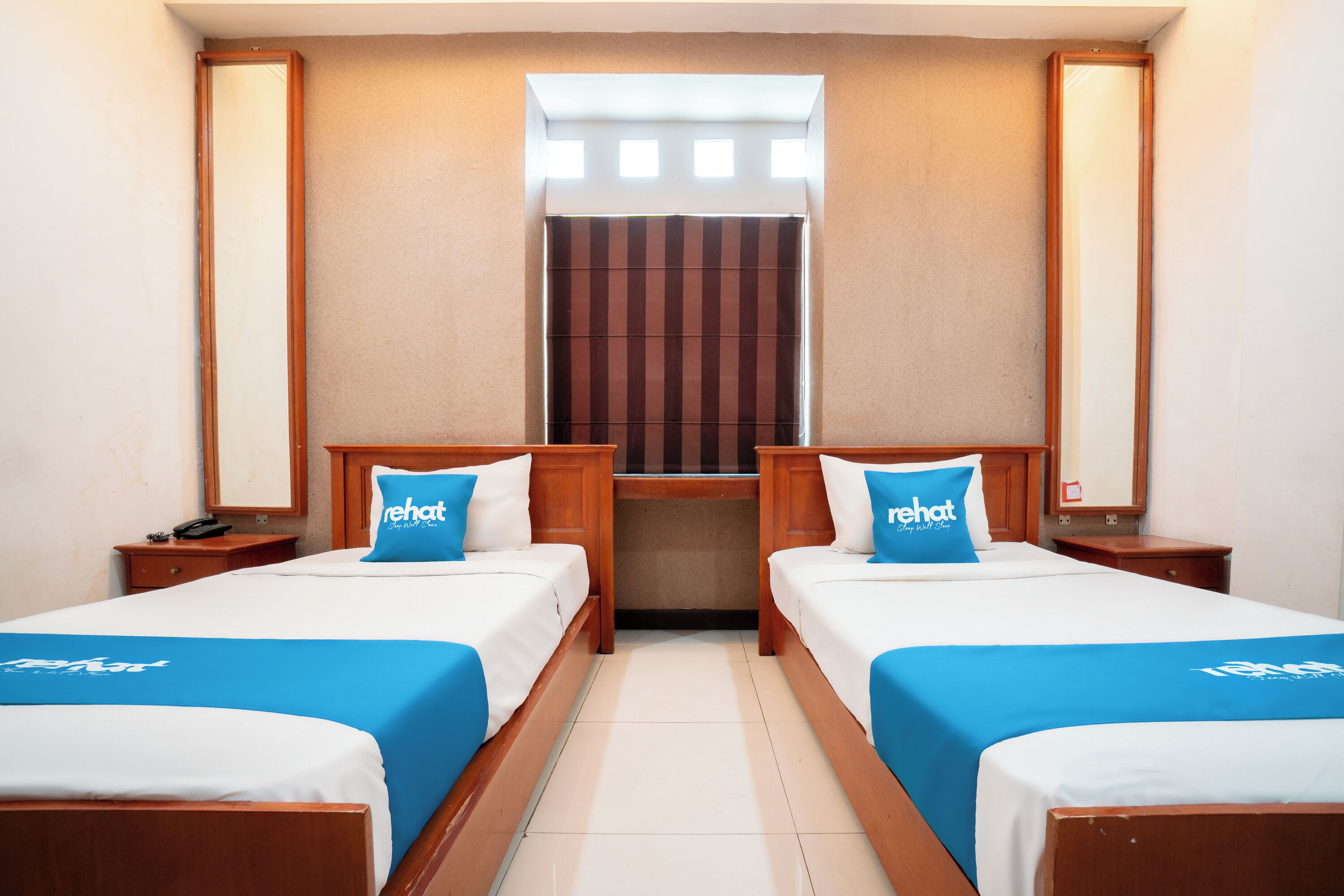Ein Hotelzimmer mit zwei Betten, jeweils mit einer blauen Tagesdecke und einem Kissen mit der Aufschrift "rehat". Ein Fenster mit einem gestreiften Vorhang, ein Holztisch und ein Nachttisch mit Telefon.