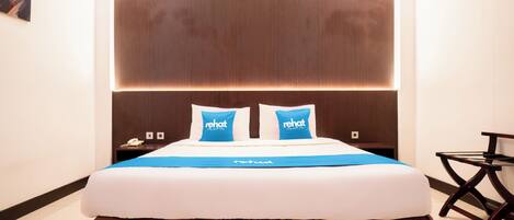 Habitación de hotel con una cama grande, dos almohadas con la marca 'rehat', una mesita de noche con un teléfono y una pequeña mesa con una silla.