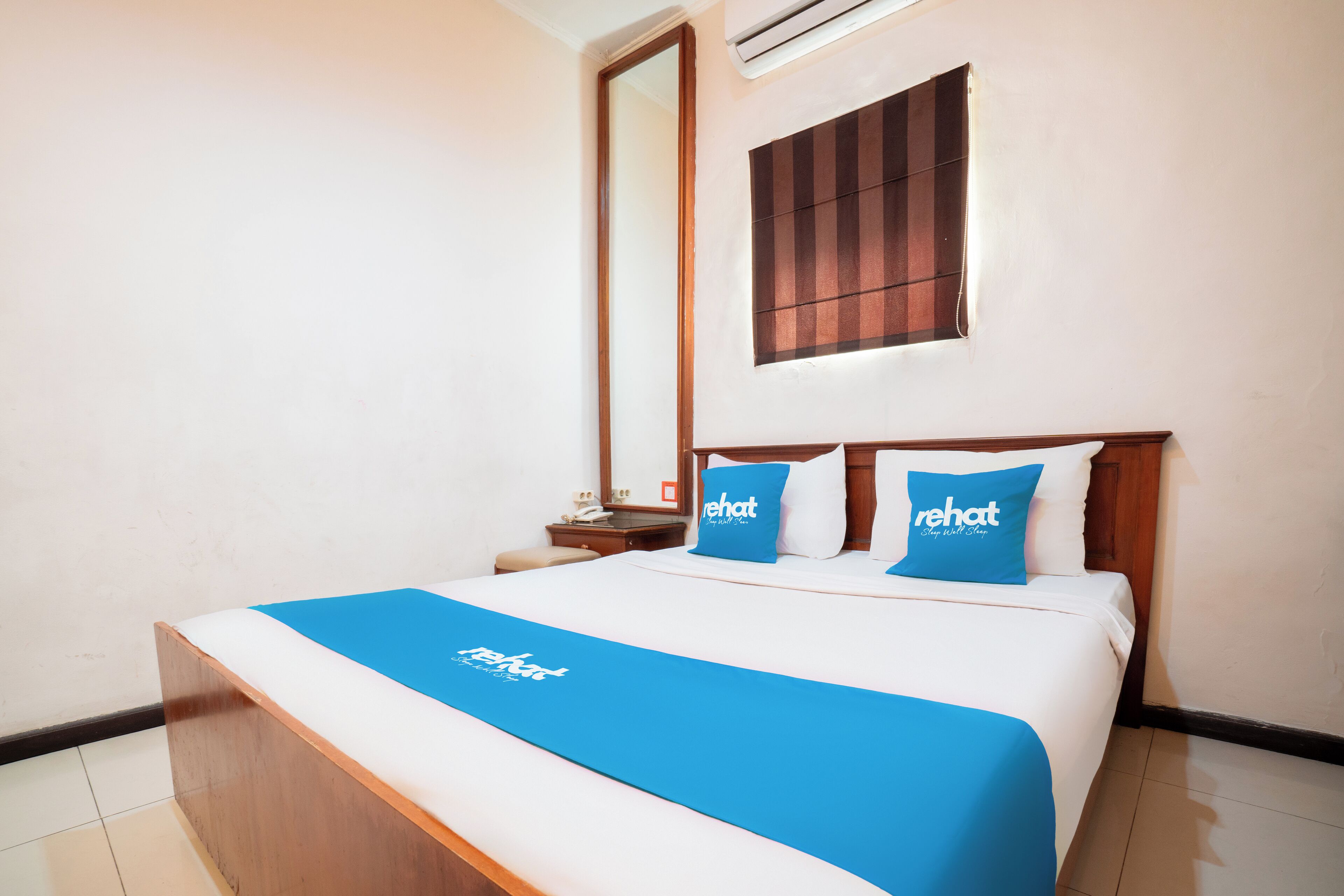 A hotel room with a bed, a nightstand, a mirror, and a window with blinds.