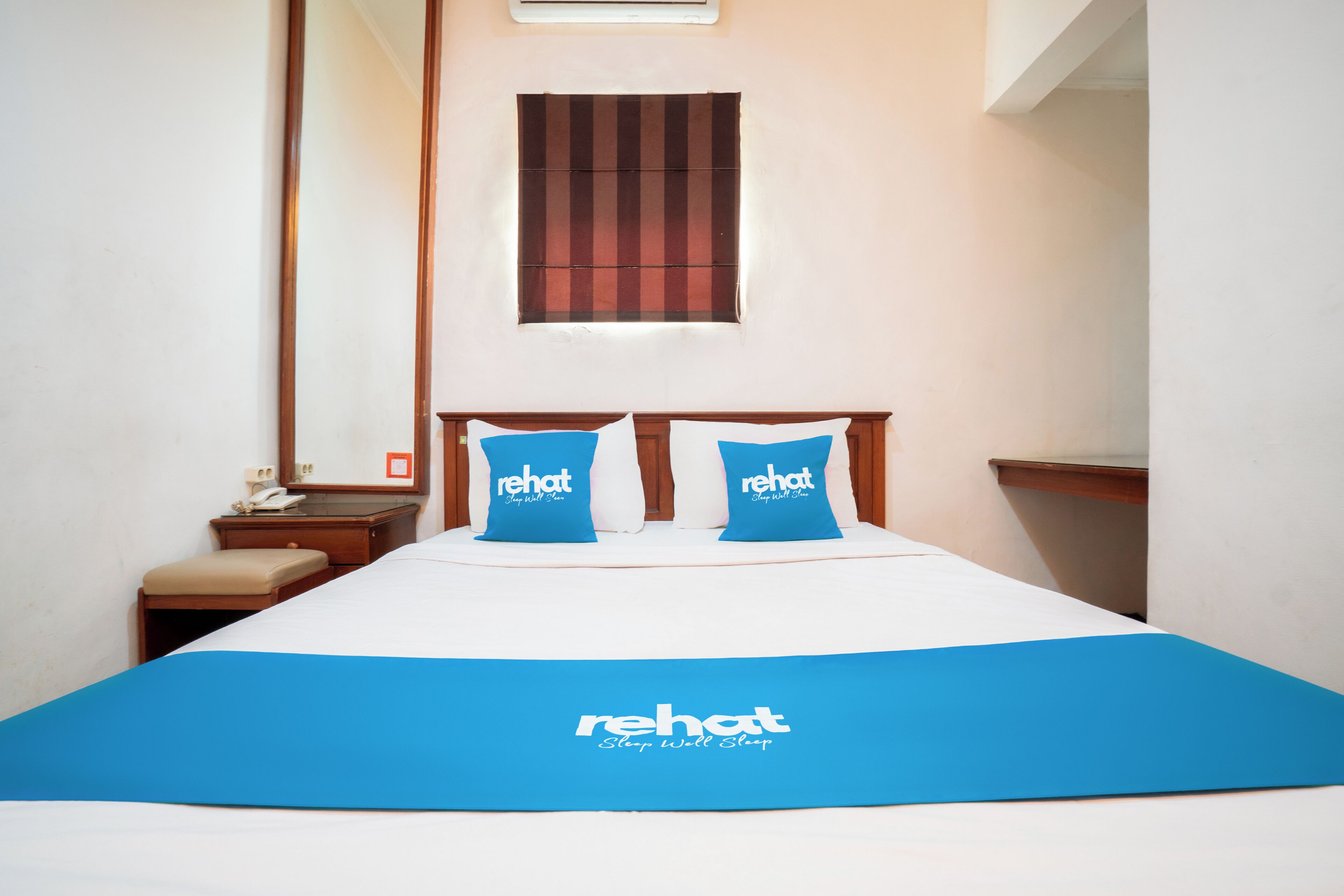 A hotel room with a bed, two blue pillows with "rehat" branding, a wooden headboard, a nightstand with a phone, and a window with blinds.