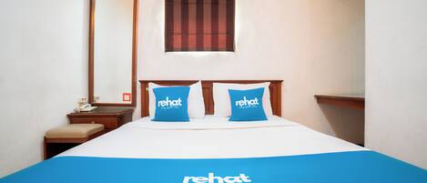 A hotel room with a bed, two blue pillows with "rehat" branding, a wooden headboard, a nightstand with a phone, and a window with blinds.