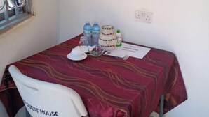 Uma pequena mesa com toalha vermelha, uma cadeira branca com o texto "GUEST HOUSE" e diversos objetos, incluindo garrafas, uma tigela e um papel com "WIFI PASSWORD" visível.