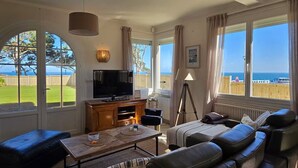 A living room with a TV, sofa, and coffee table, large windows with a view of the sea, and a lamp. - "Les pieds dans l'eau": panoramic sea views and direct beach access! (Saint-Quay-Portrieux)