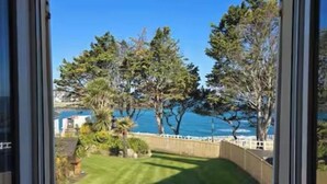A view through a window of a grassy yard with trees and a body of water in the background. - "Les pieds dans l'eau": panoramic sea views and direct beach access! (Saint-Quay-Portrieux)