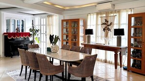 Uma sala de jantar ampla, com uma mesa grande e cadeiras de vime, sobre um tapete estampado.