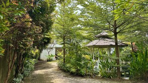 Caminho no jardim que leva a um gazebo, cercado por vegetação exuberante e um edifício branco ao fundo.