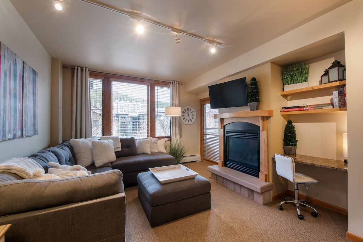A modern living room with a sofa, ottoman, fireplace, television, and bookshelves.