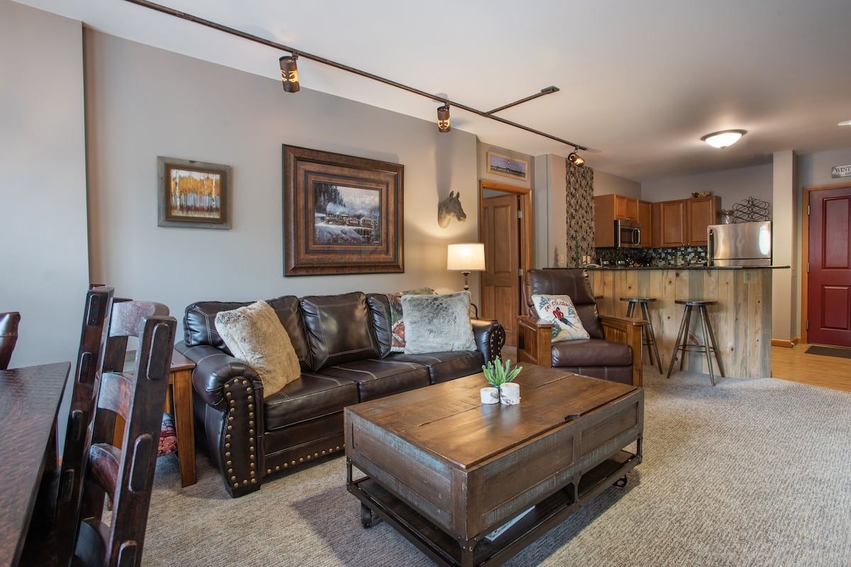 A living room with a dark brown leather sofa, a wooden coffee table, and a dining table with chairs. There is a kitchen area with wooden cabinets and a red door.