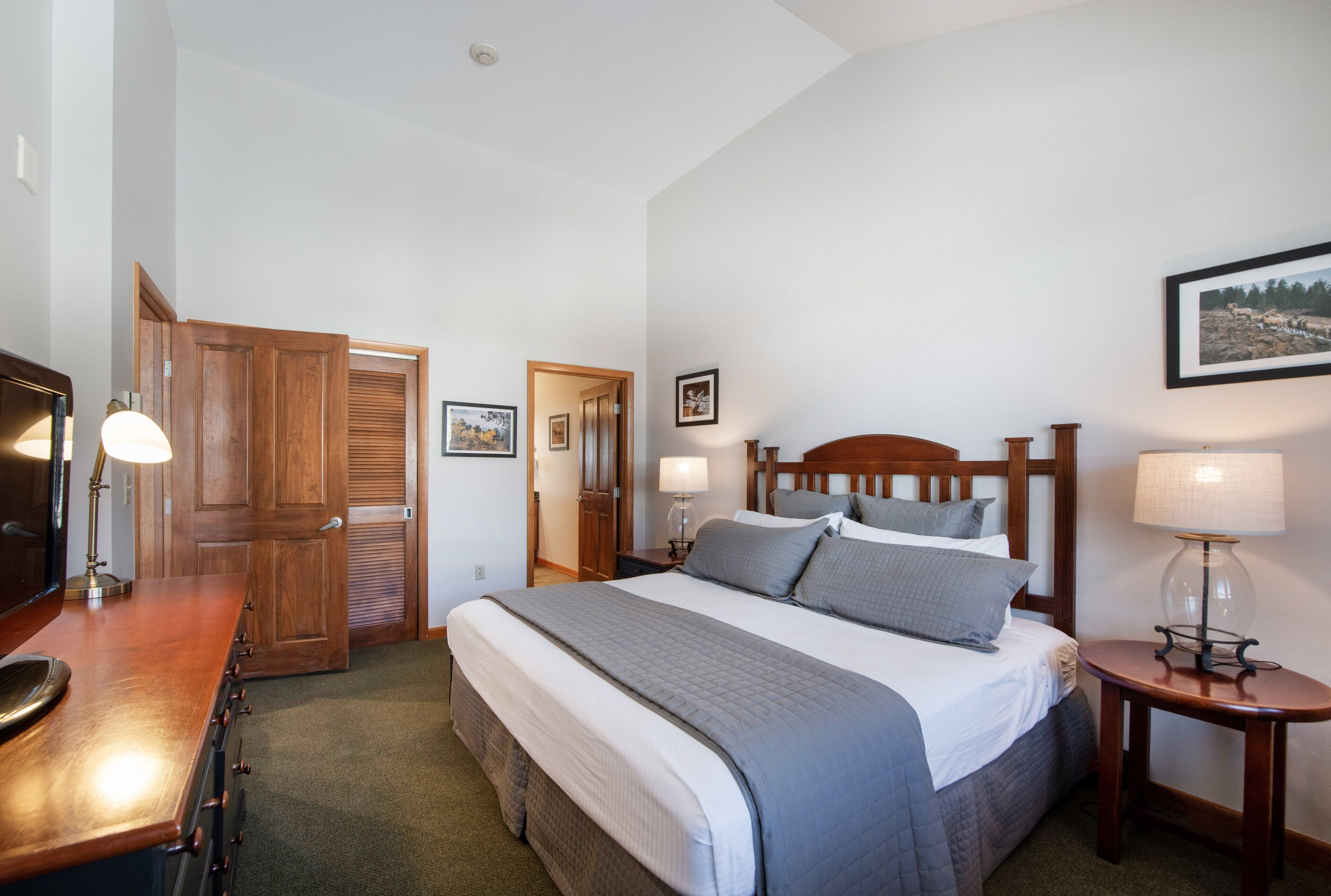 A hotel room with a wooden bed, a desk with a lamp, a bedside table with a lamp, and a framed picture on the wall.