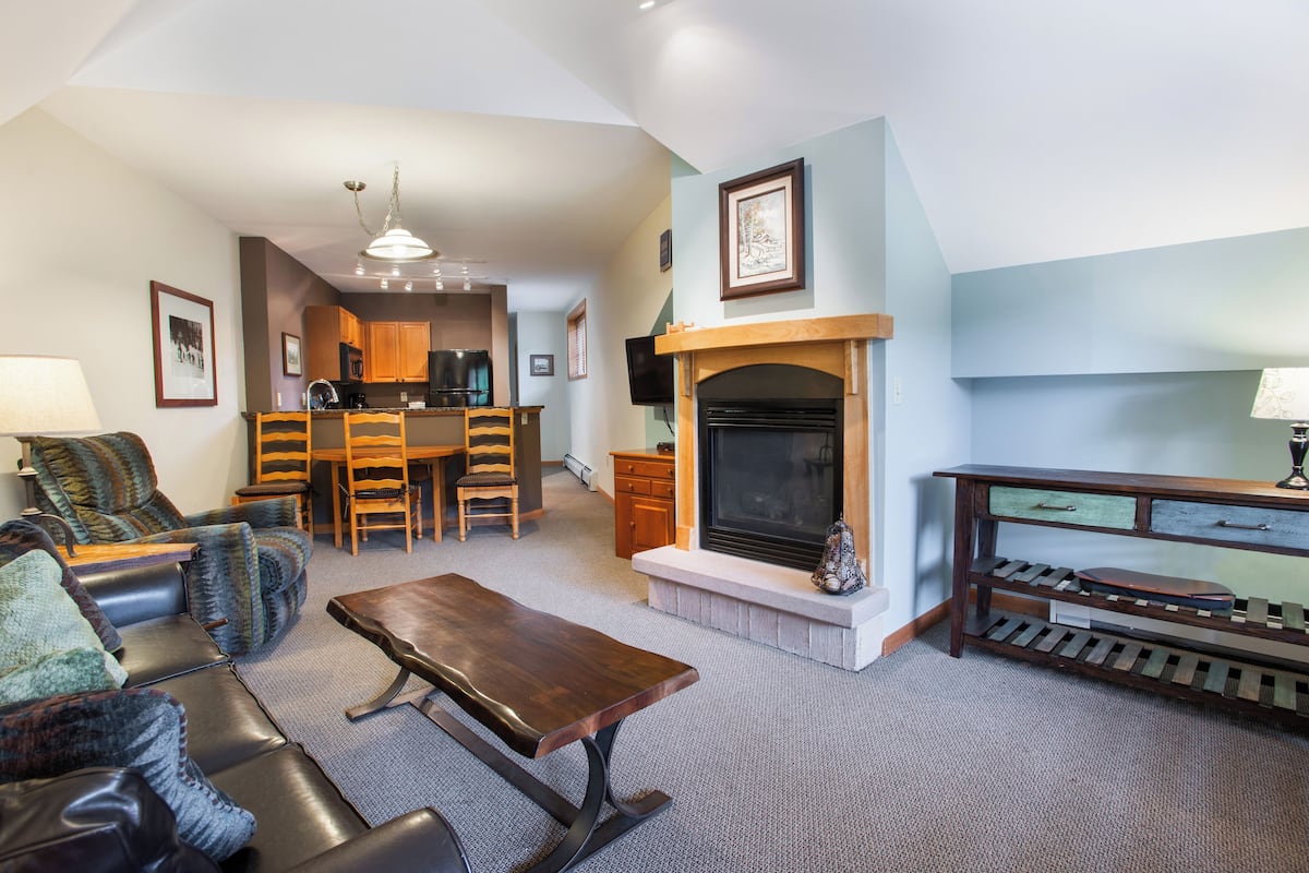 A spacious living room with a fireplace, a dining area, and a kitchen in the background.