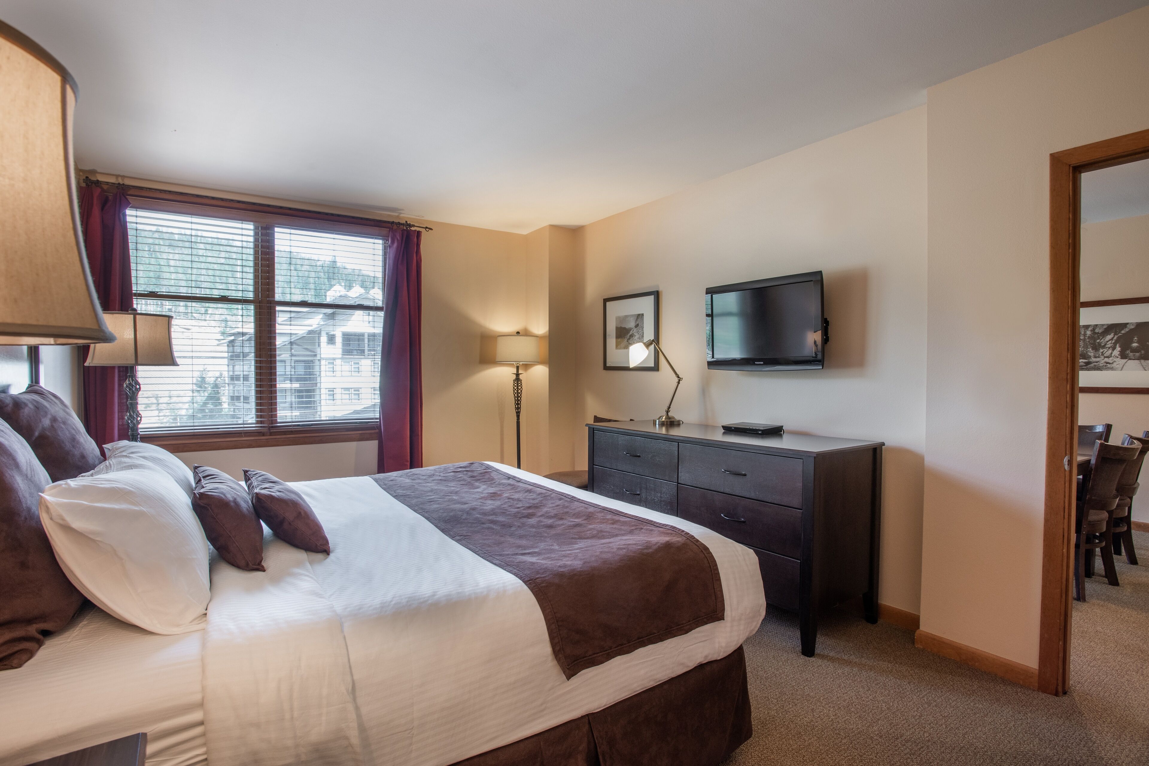 A hotel room with a large bed, a desk, a chair, a TV, and a window with blinds.