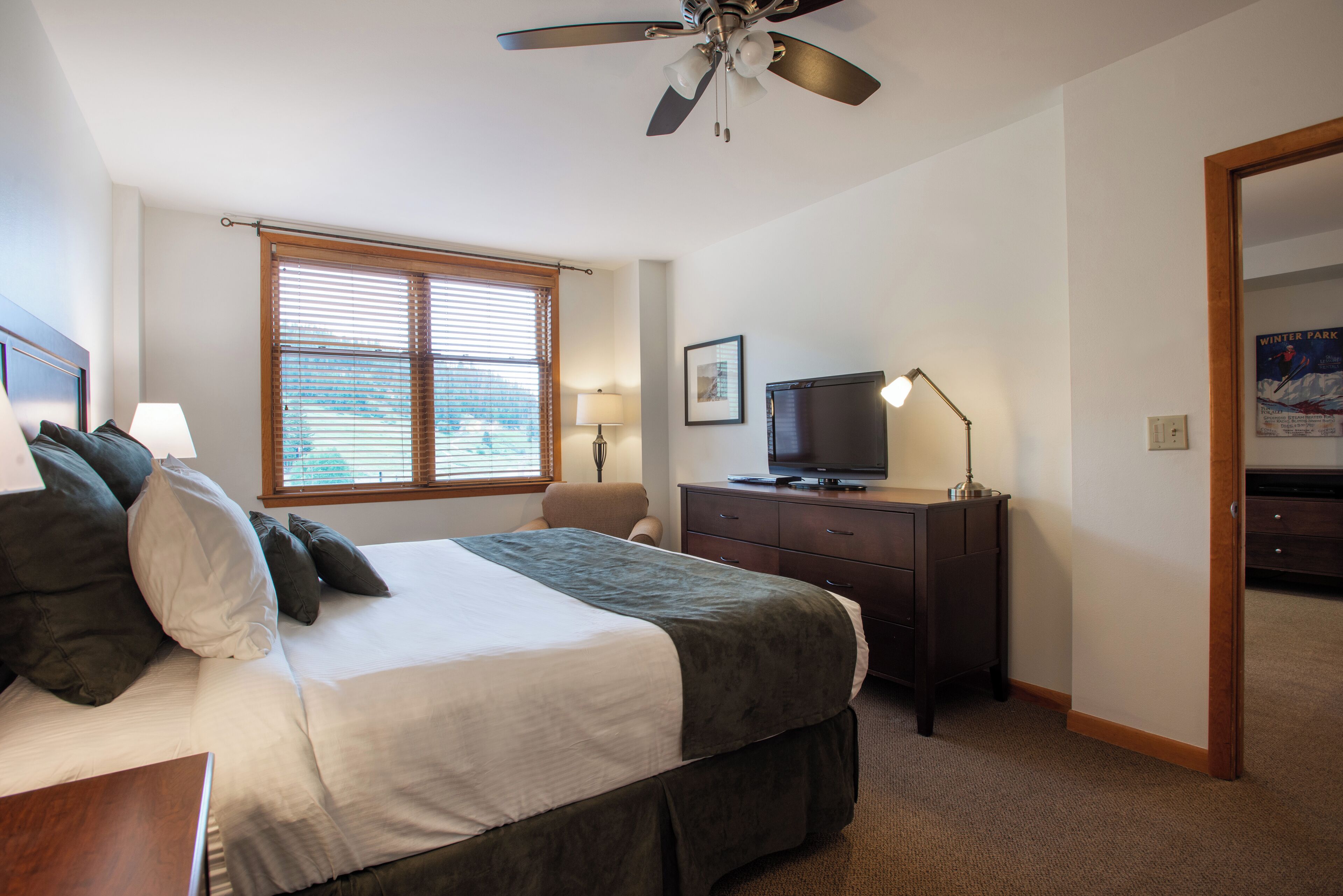 Ein Hotelzimmer mit Bett, Schreibtisch, Fernseher, Deckenventilator und einem Fenster mit Jalousien.