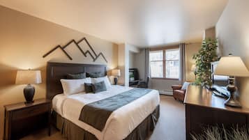 A hotel room with a large bed, two bedside tables with lamps, a desk with a chair, a potted plant, and a window with curtains.