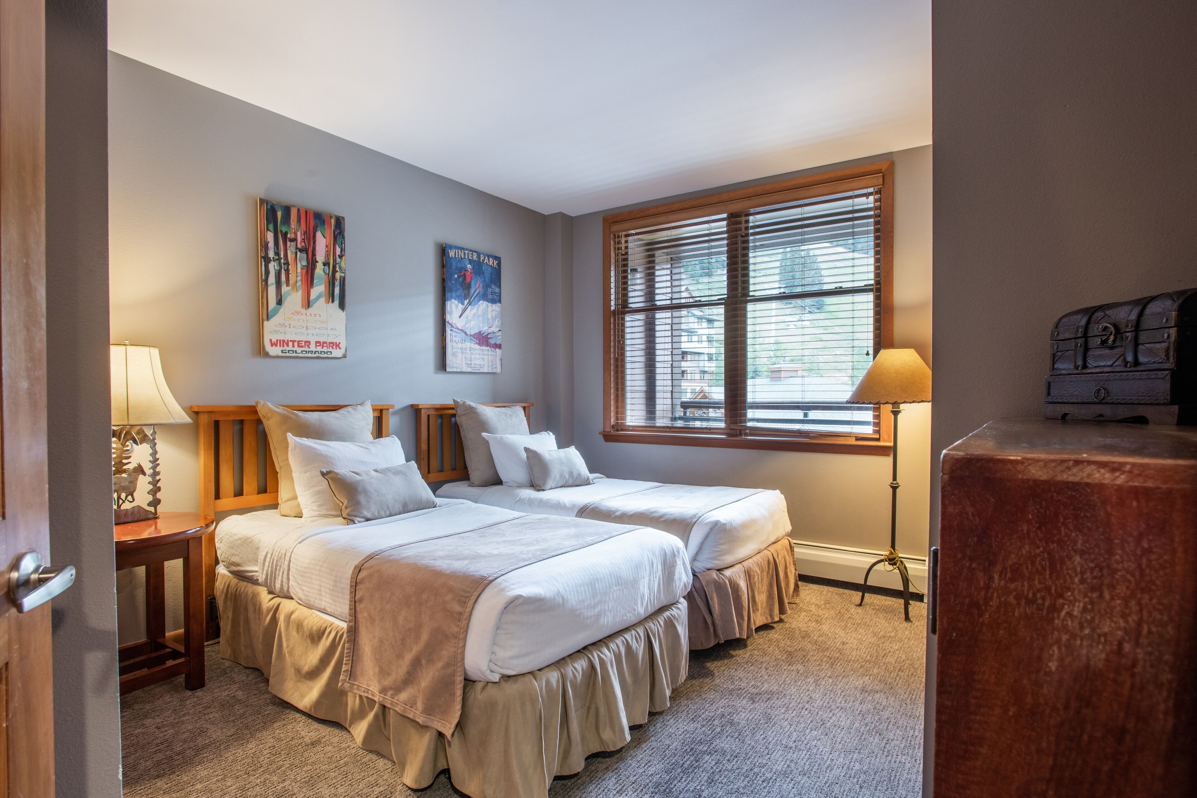 Une chambre avec deux lits, une fenêtre avec des stores, une lampe et une valise ancienne.
